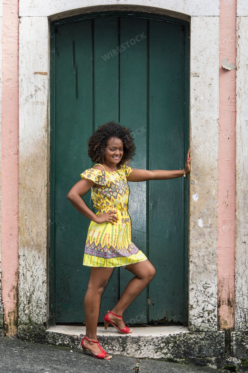 An African female model with curly black hair and yellow dress posing next to a green door in Brazil