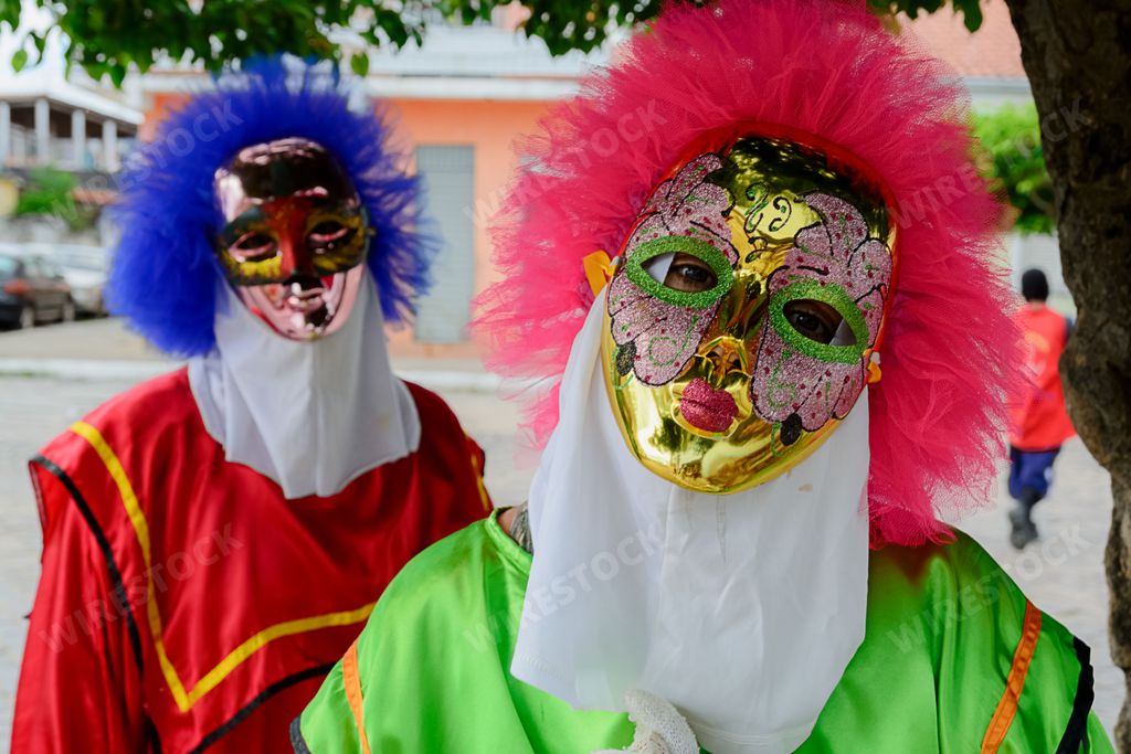 A group of people wearing colorful clown masks and costumes are seen in front of a line of trees on a city street