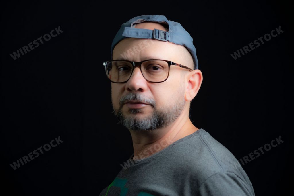 A portrait of a bald man in a cap and glasses looking at the camera against the black background