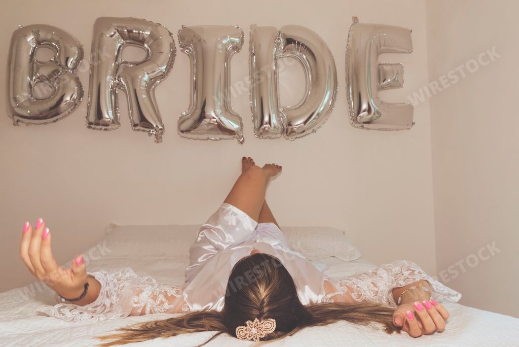 A Brazilian female lying on a bed against a white wall with silver balloons written bride