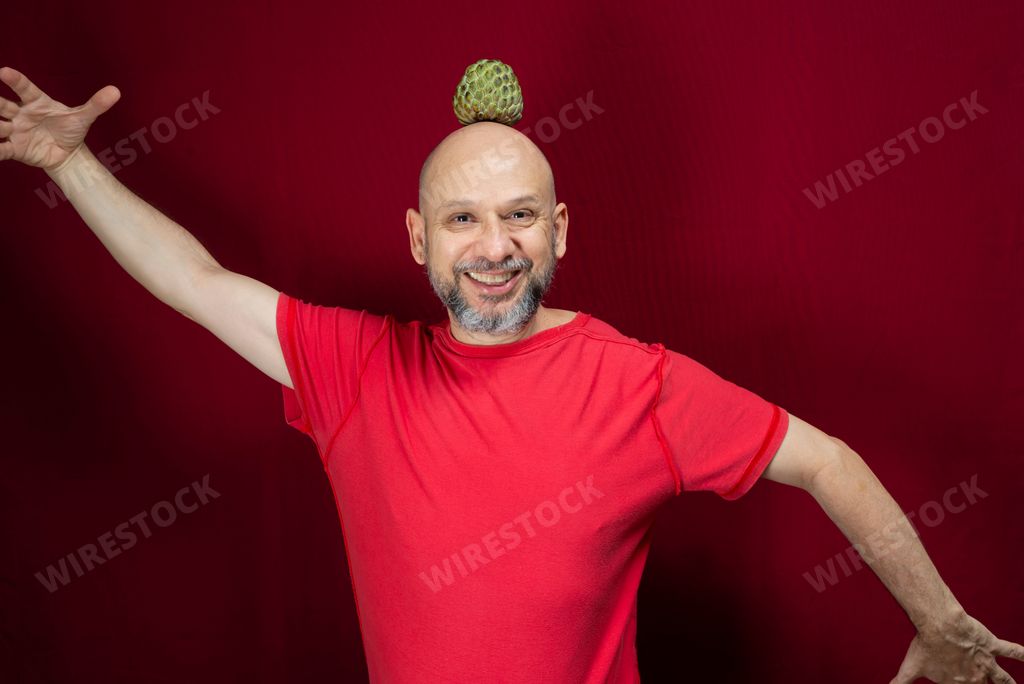 Young handsome man with beard, bald head and red shirt standing with pinecone fruit balanced on head against red background.