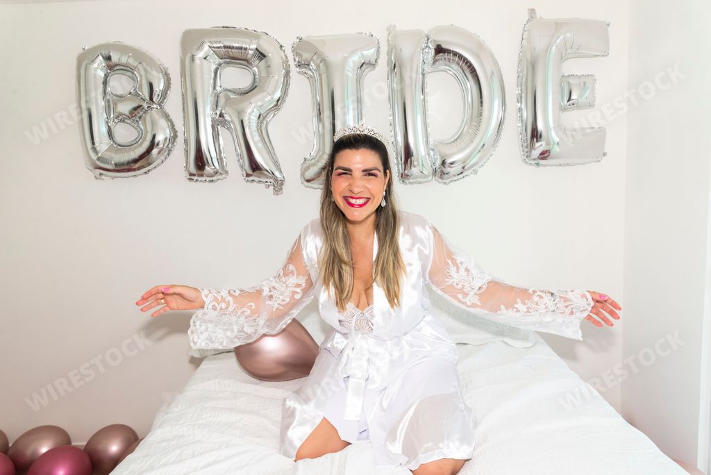 A young happy Brazilian woman in a white silk robe sitting on a bed in front of a Bride balloon at Bachelorette party