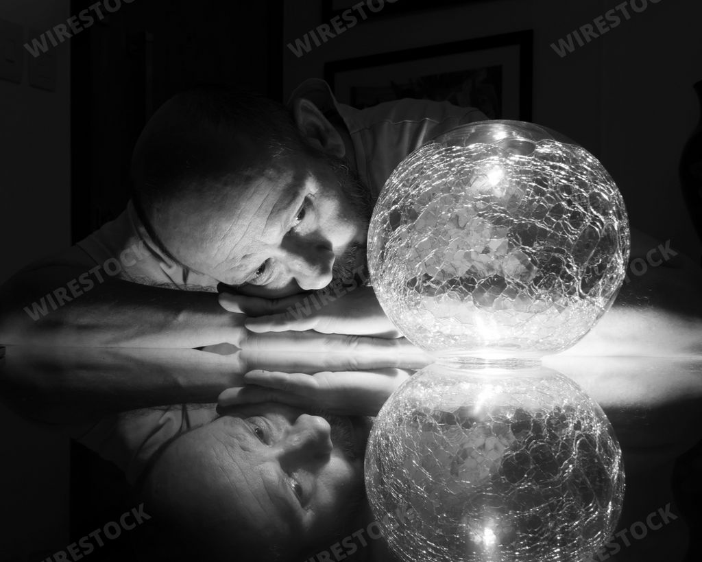 A grayscale closeup of a man in the darkness leaning on the table and looking at a glass ball.