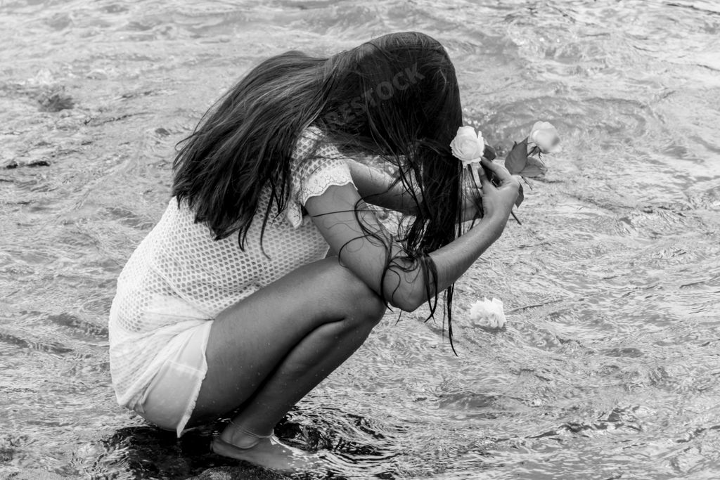 A high-contrast black and white image of a woman kneeling in shallow water, her hands cupping several small pebbles