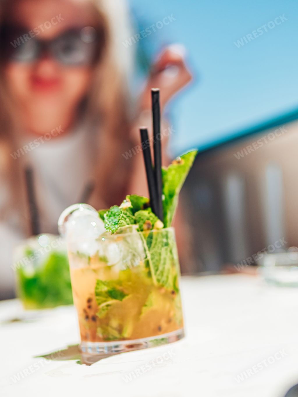 A selective focus shot of two glasses of mojitos and a female with sunglasses sitting at the white table