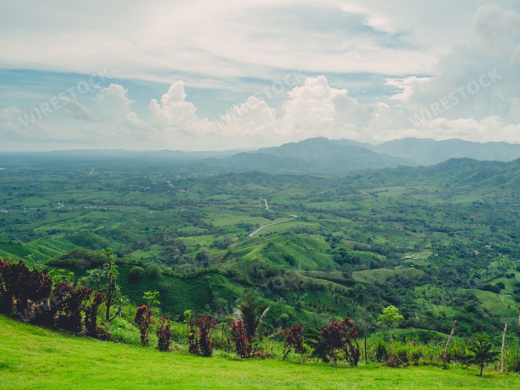 A landscape of hills covered in greenery with mountains on the background under a cloudy sky