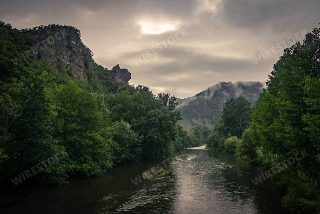 A river surrounded by rocks covered in mosses and forests under the sunlight and a cloudy sky