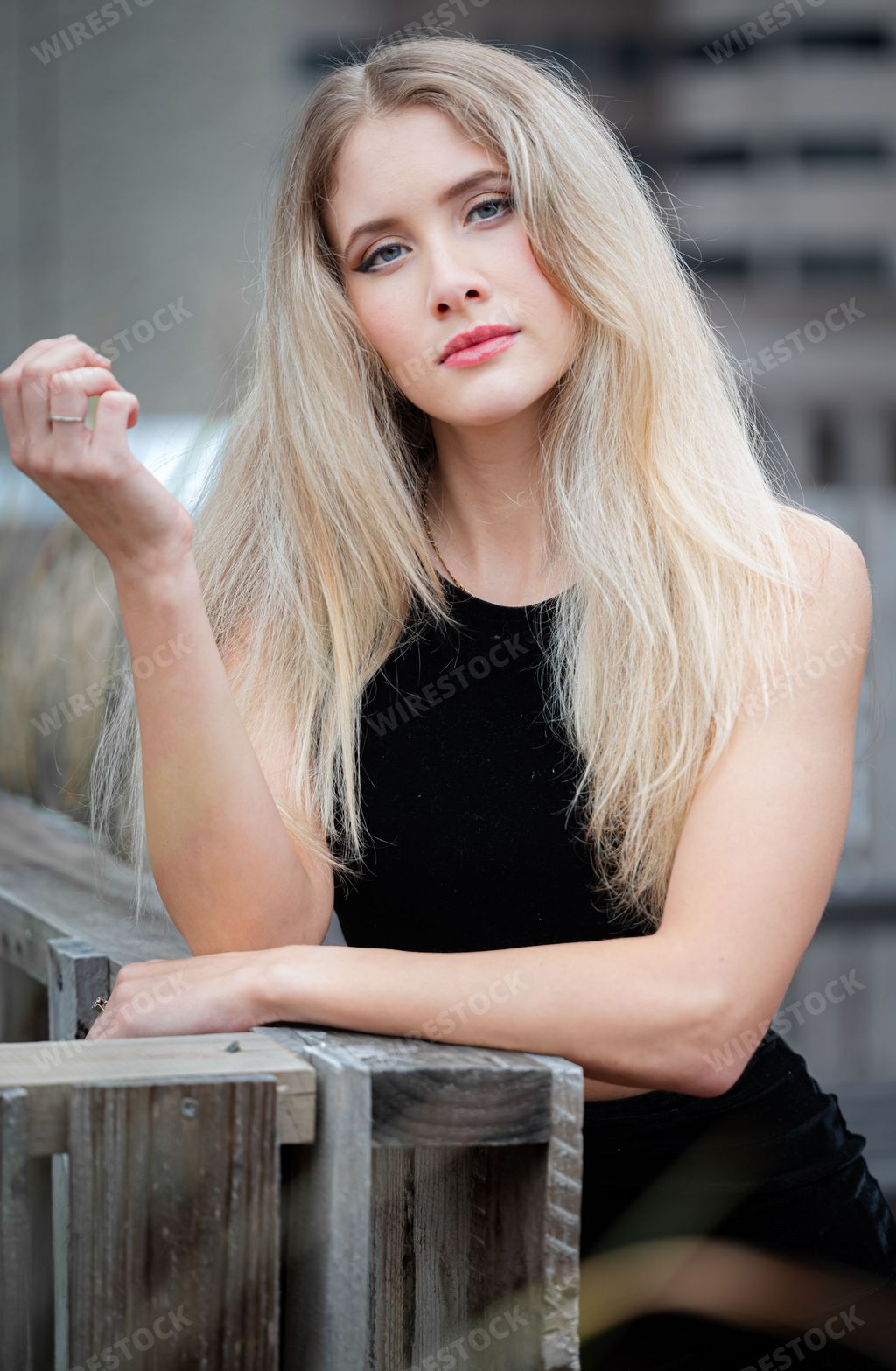 A blonde girl wearing a black evening dress leaning on a wooden railing in an urban setting