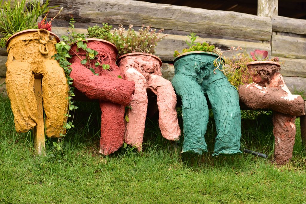 An outdoor scene featuring five brightly-colored flower planters nestled in a grassy area next to a wooden fence