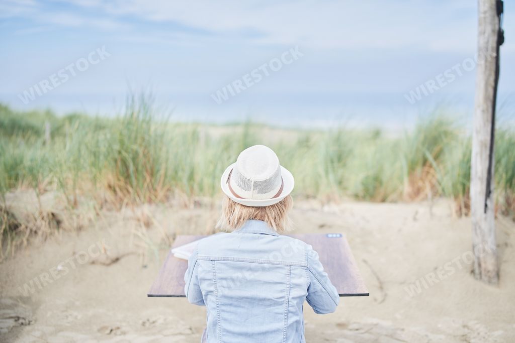 A back view of an unrecognizable blonde woman working on a laptop at a beach in summer
