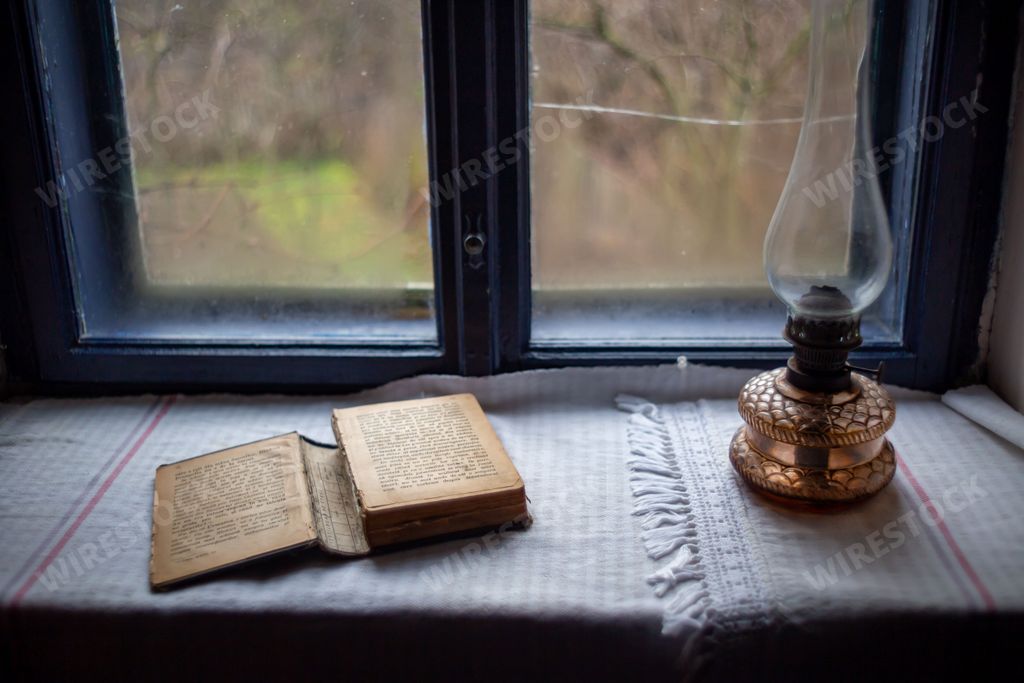 An old open book resting on a wooden table next to a white lamp, illuminated by the warm yellow light of the bulb