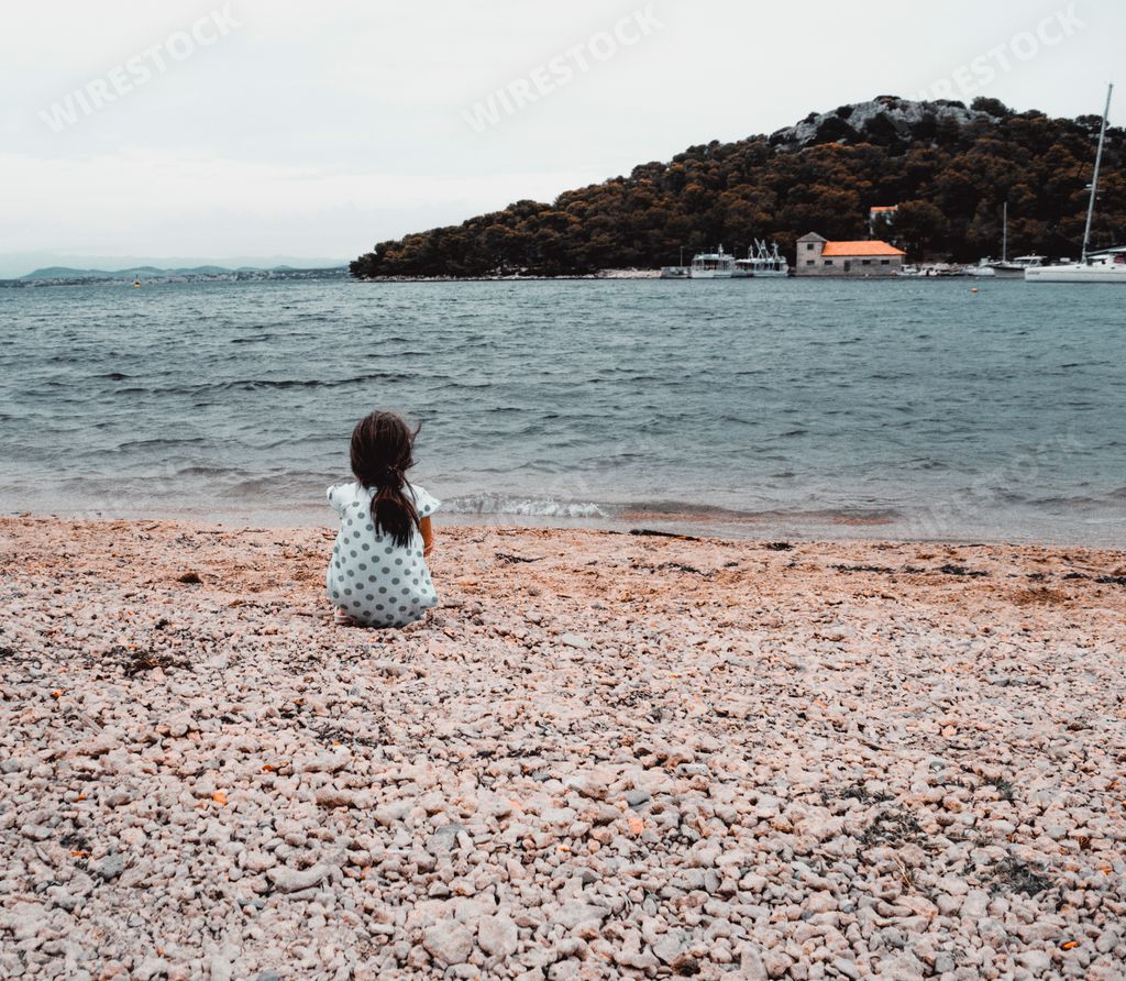 A vertical shot of a little girl sitting alone on the beach looking at the blue sea