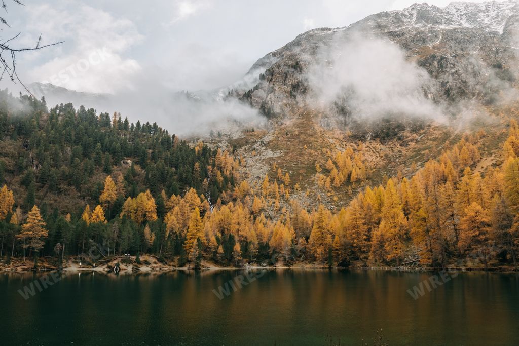 A calm reflective lake and low flying clouds covering a rough mountain covered with colorful autumn foliage