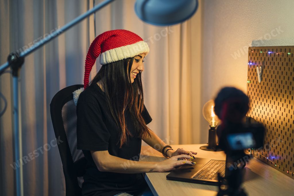 A charming female blogger with a laptop and a knitted Santa hat recording a video tutorial  in front of a camera