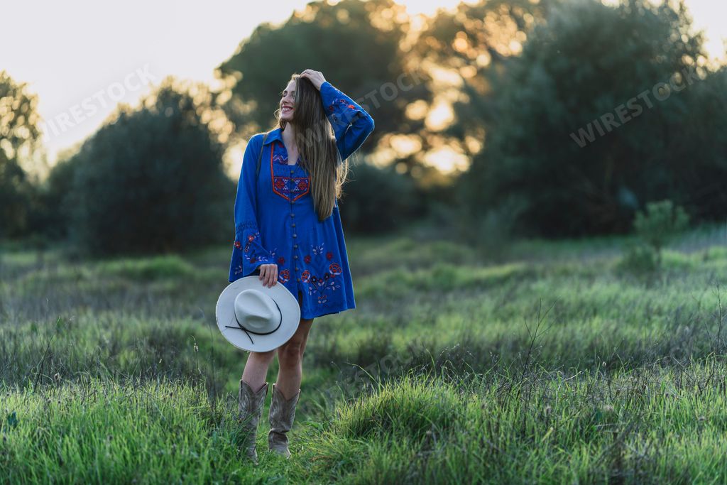 A young female adult stands in a grassy meadow, wearing a light blue dress and a sun hat in her hands