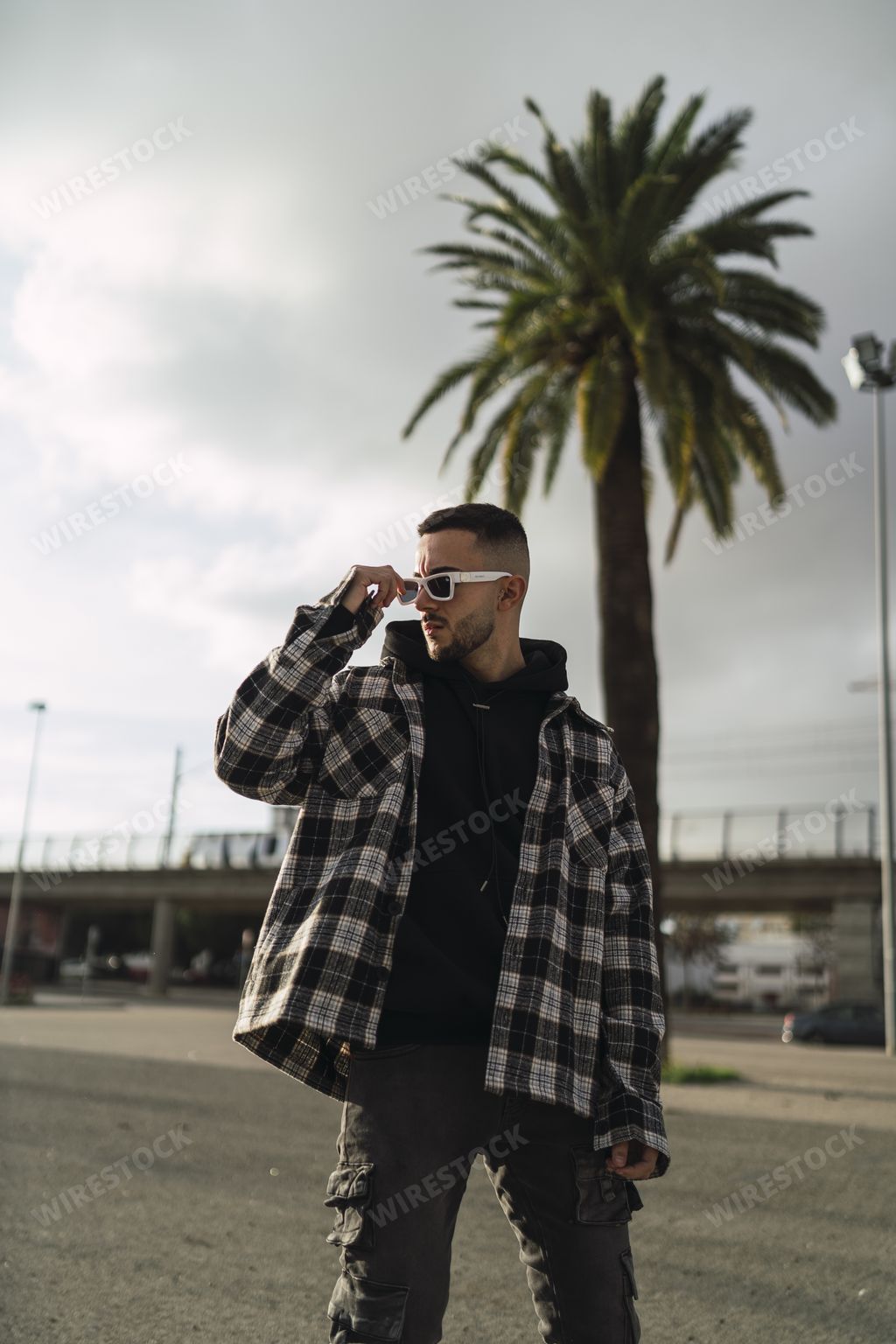 A vertical shot of a stylish guy wearing a plaid jacket, a black hoodie, black pants, and sunglasses