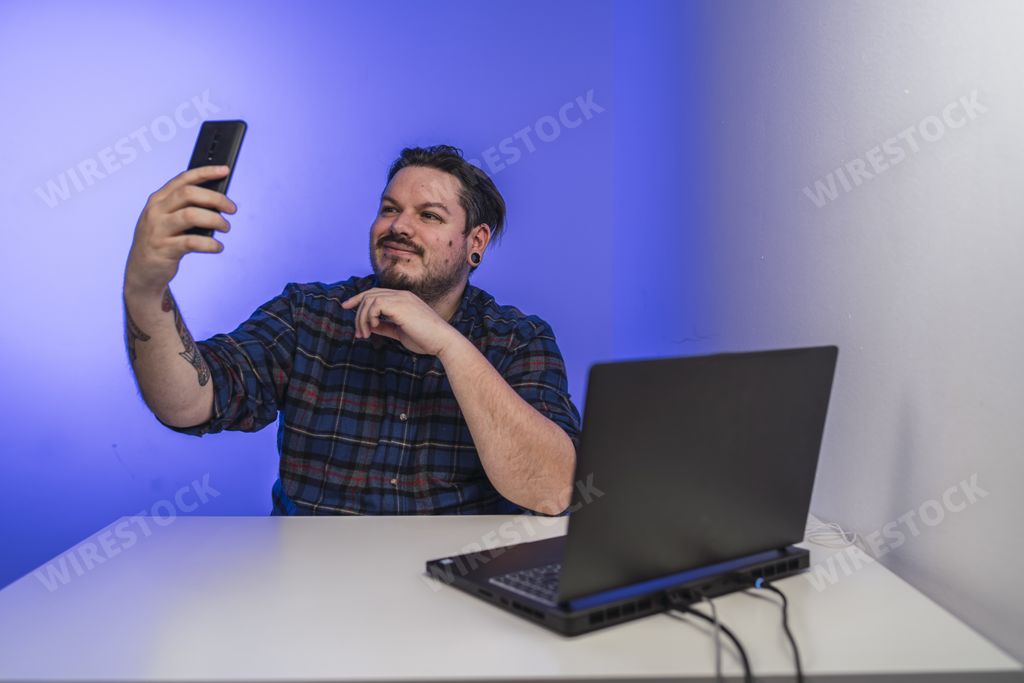 A Spanish bearded man with tattoos taking a selfie while working on a laptop