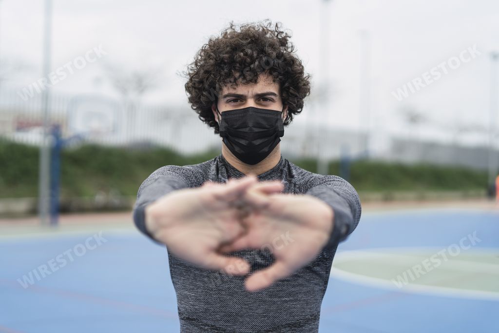 A young athletic male wearing a black mask has interlaced the fingers and stretching the arms with palms out