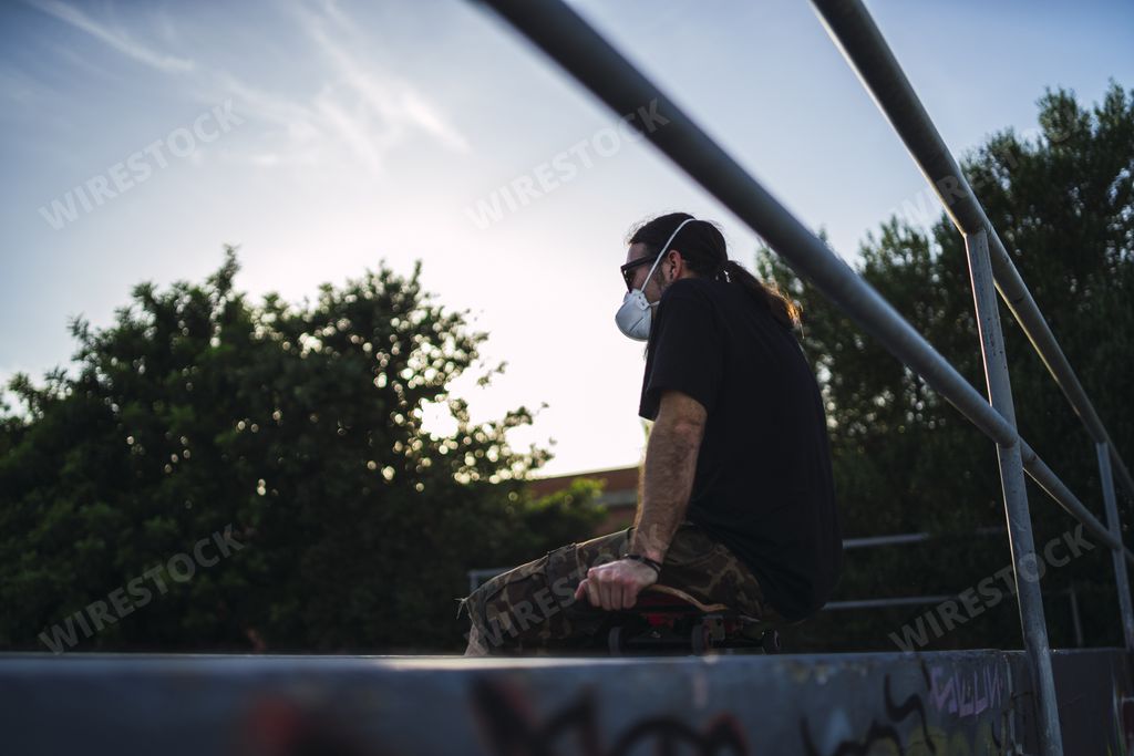 A low angle shot of a male wearing a face mask sitting on his skateboard at the park- Covid-19 concept