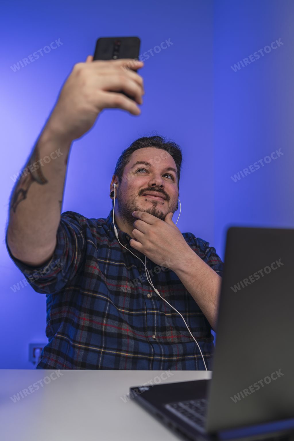 A bearded Spanish man with earphones taking a selfie while working on a laptop