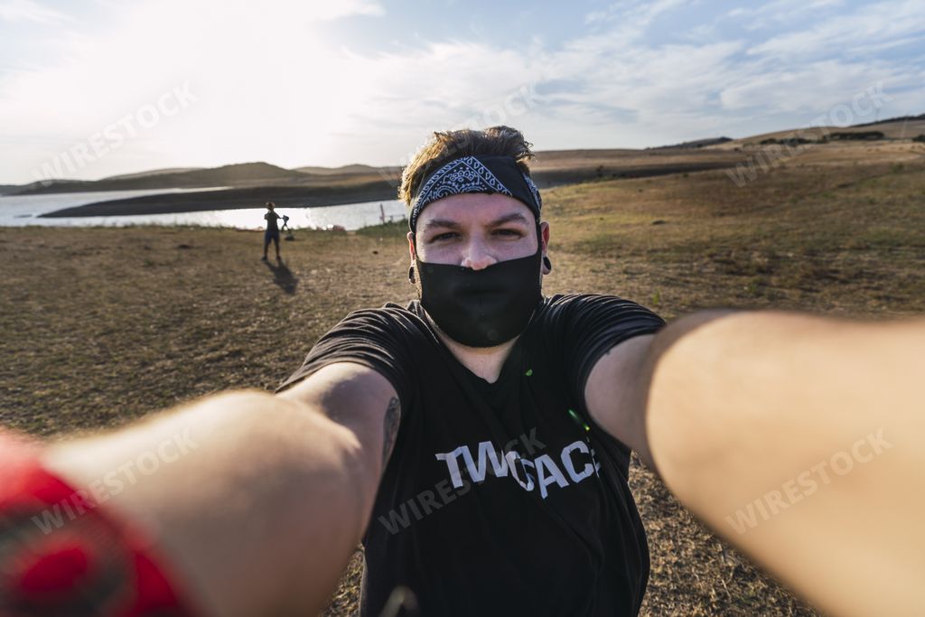 A male wearing a mask taking a selfie near a lake