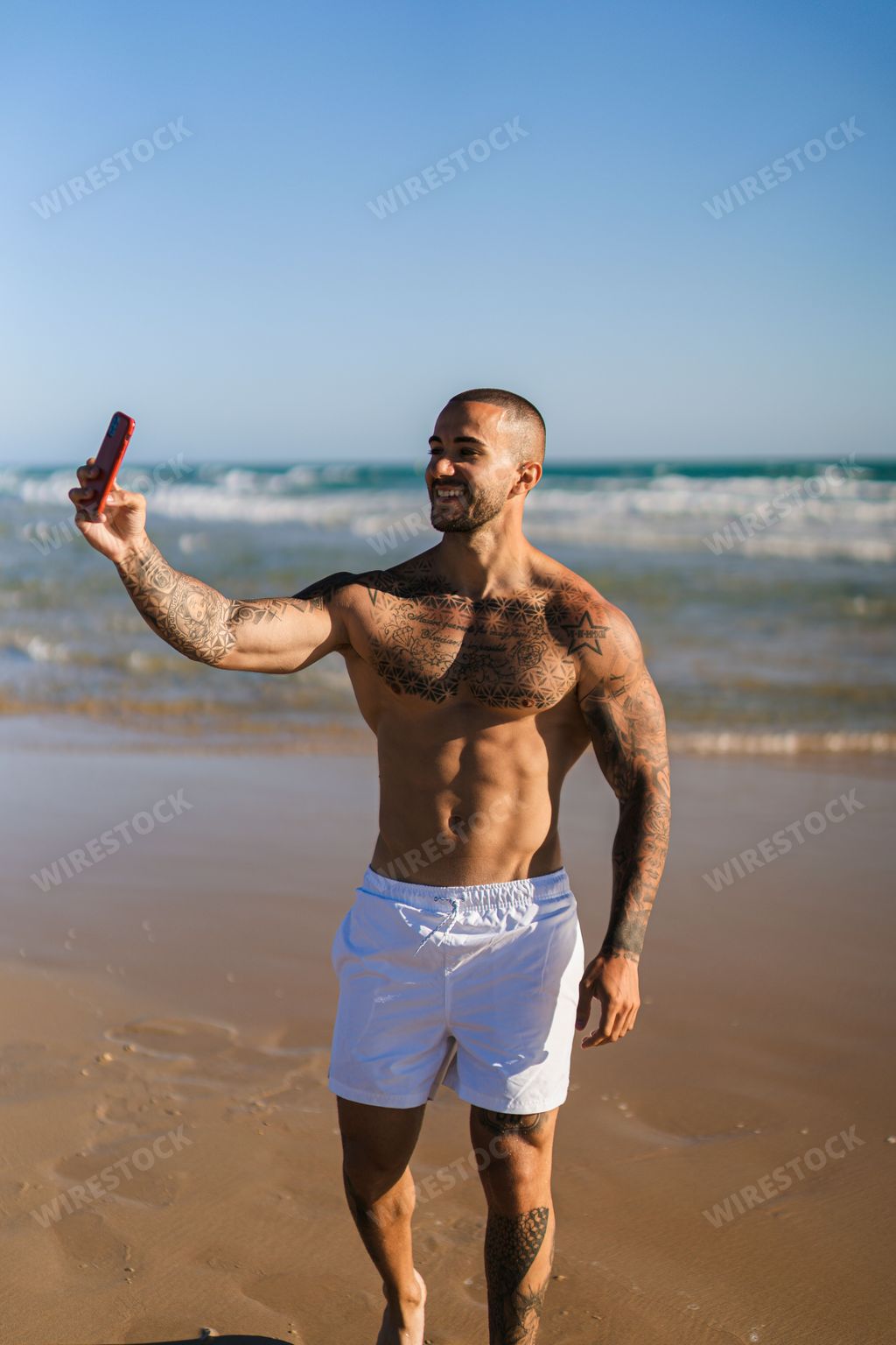 A vertical shot of an attractive topless man with tattoos standing  on a beach and taking selfies with his phone