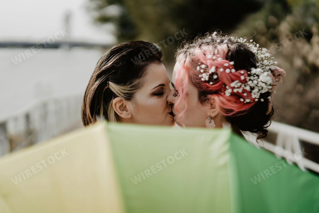 A closeup of a lesbian pair kissing each other and holding a rainbow umbrella on the bridge