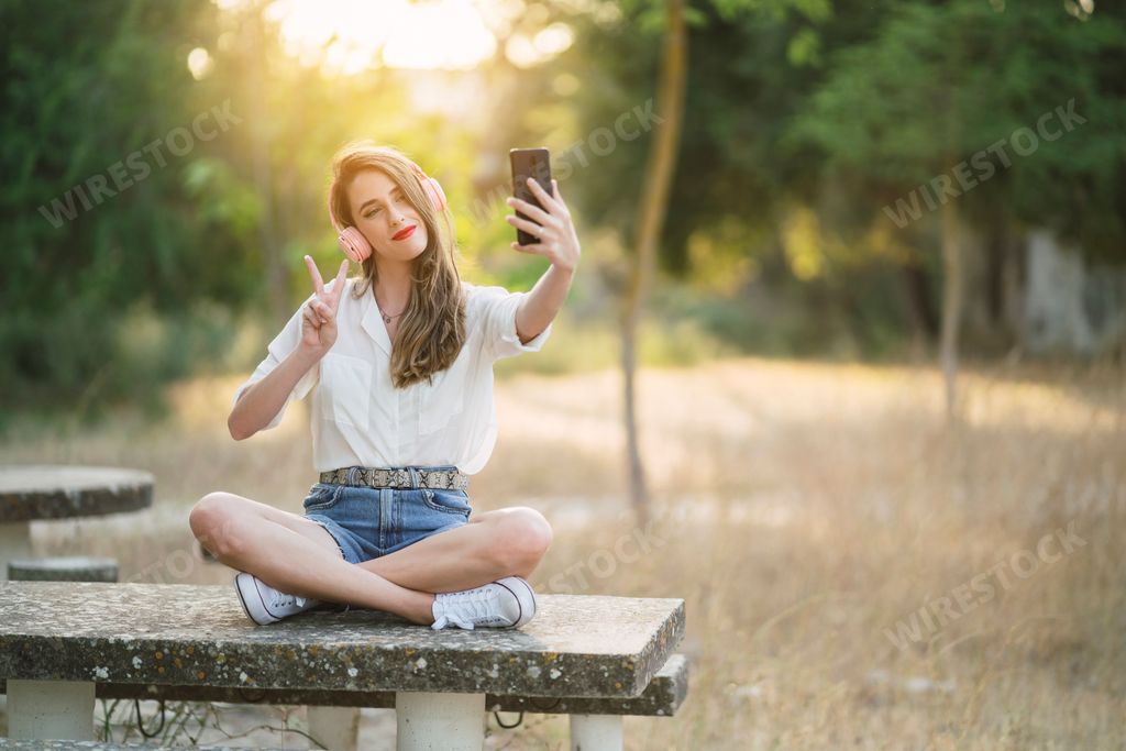 A young Spanish woman wearing shorts with a shirt and taking a selfie with a phone in a park