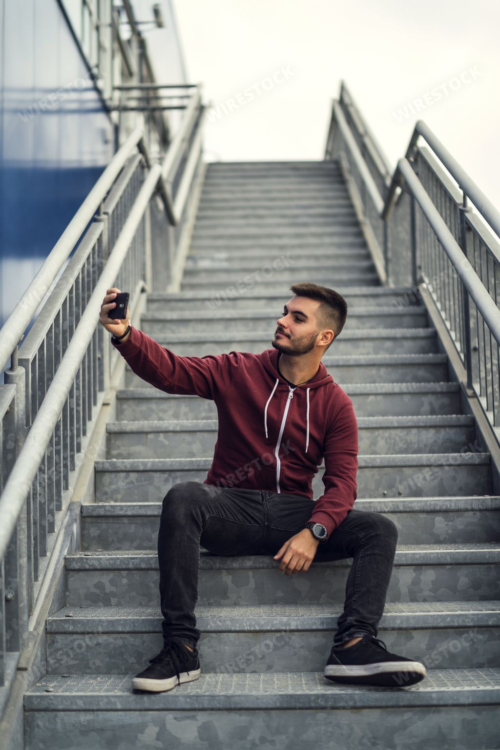 A young male wearing a red jacket with dark jeans and taking a selfie with a phone while sitting on the stairs outdoors