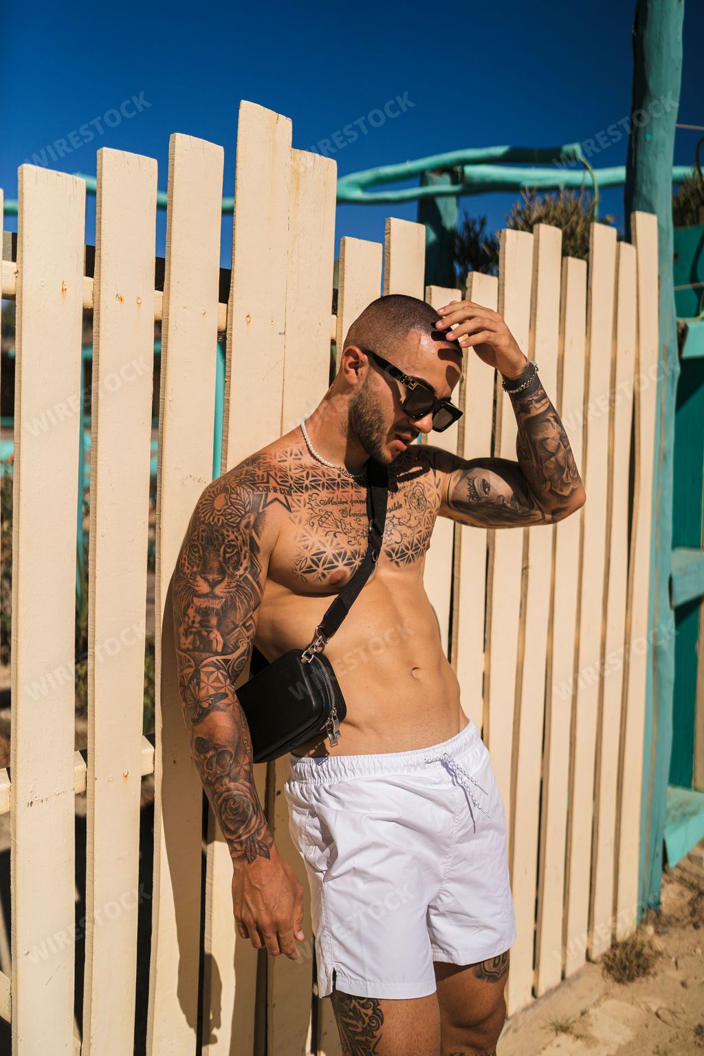 A vertical shot of an attractive topless man with tattoos standing next to a wooden fence with a blue sky