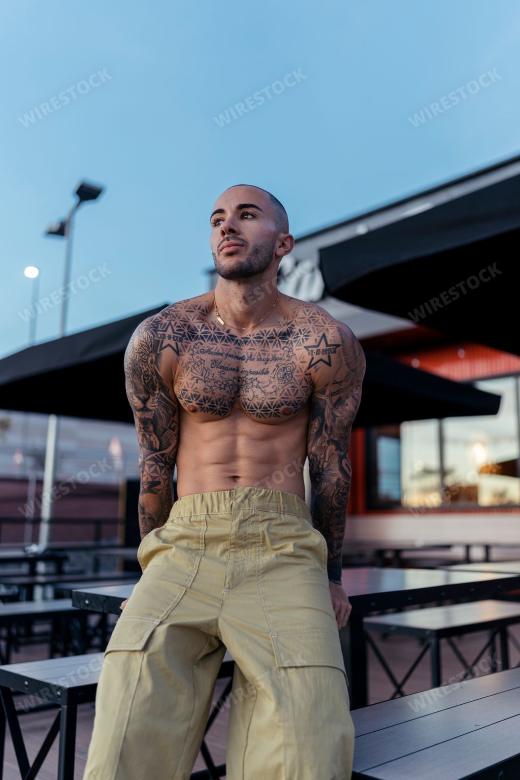 A handsome shirtless man with tattoos, posing against an outdoor restaurant