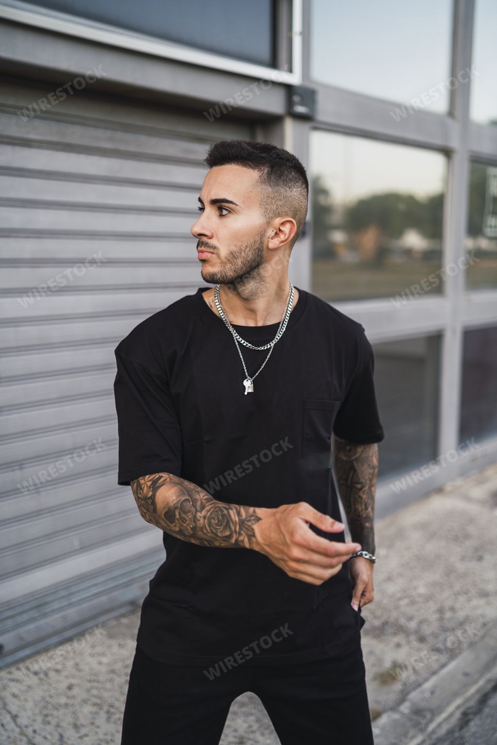 A vertical shot of a young European tattooed hip male in a black sporty outfit and a metal chain standing in front of a garage