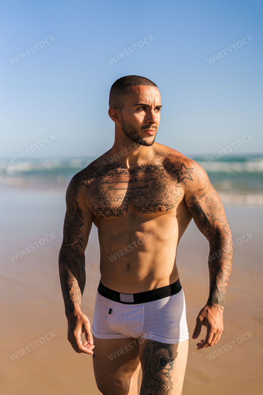 A vertical shot of an attractive fit tattooed guy at a sandy beach on a beautiful summer day