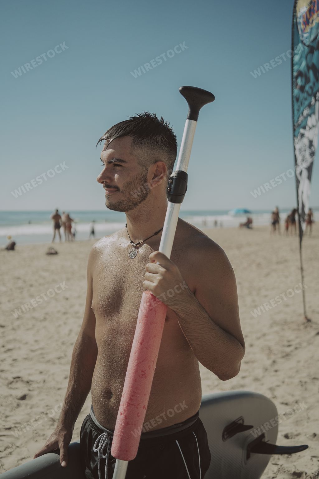 A vertical shot of a young attractive shirtless male holding a paddle and a surfboard at the beach