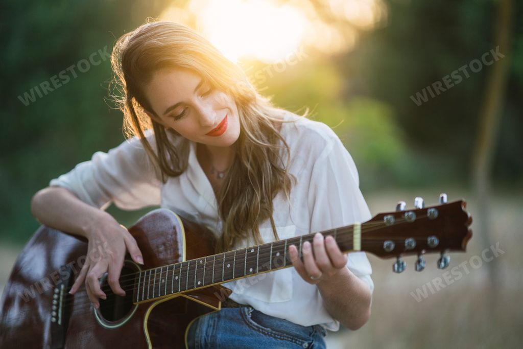 A young Spanish woman sitting in a park and playing the acoustic guitar on a sunny day