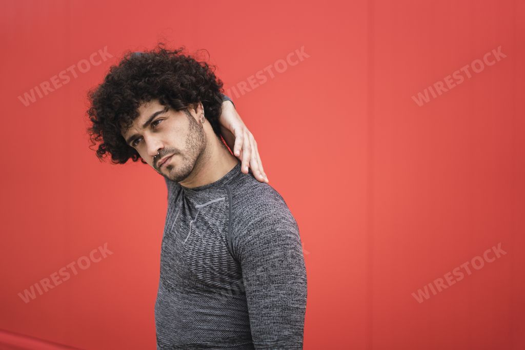 An attractive Spanish guy posing by the red wall with one hand behind the neck; background with a copy space