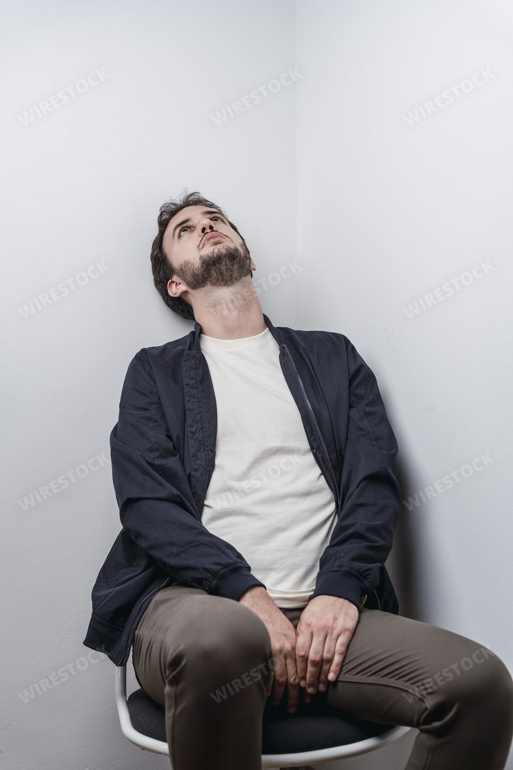 A vertical shot of a Caucasian man staring at the ceiling while seated on a chair at a corner of a room