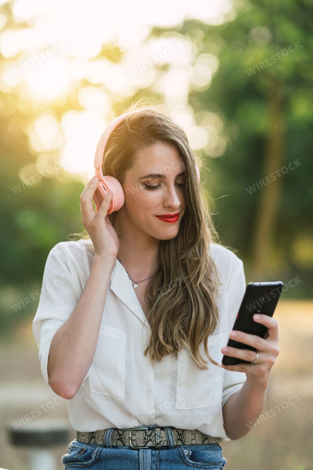 A young Spanish woman listening to music with pink headphones and a phone while sitting in a park