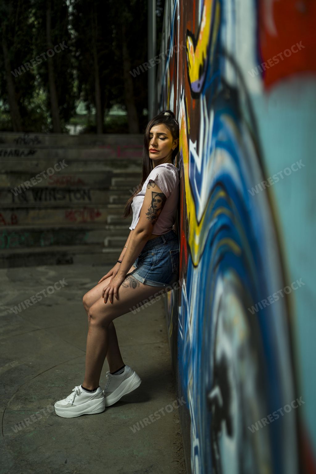 A vertical shot of a young caucasian tattooed female posing while leaning on a wall with colorful graffitis