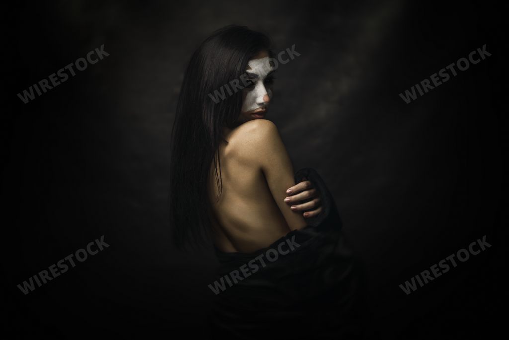 A half-naked female wearing clown makeup on her face standing in front of a black background