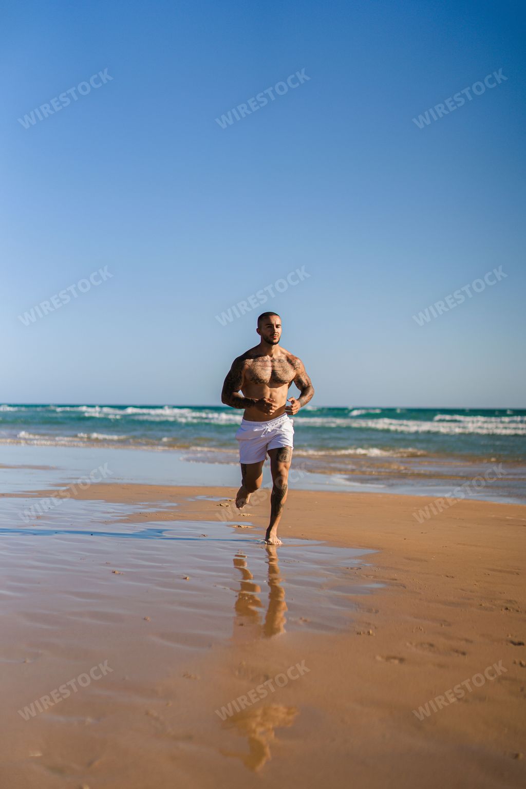 A vertical shot of an attractive topless man with tattoos running on a beach next to a sea