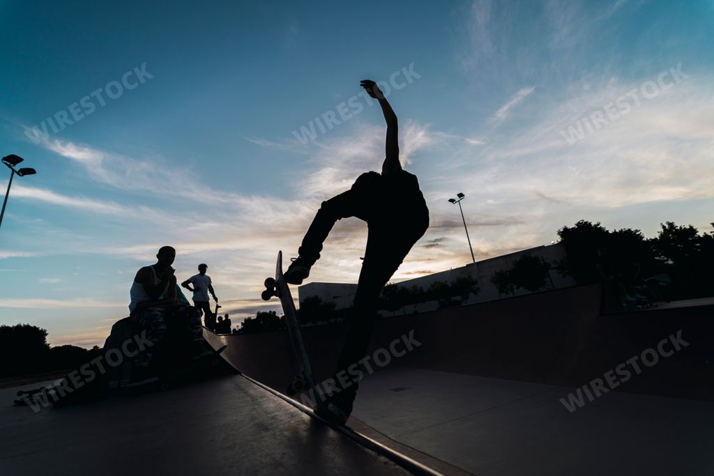 A low angle shot of a man skateboarding in a skatepark with his friends under a blue sky at sunset