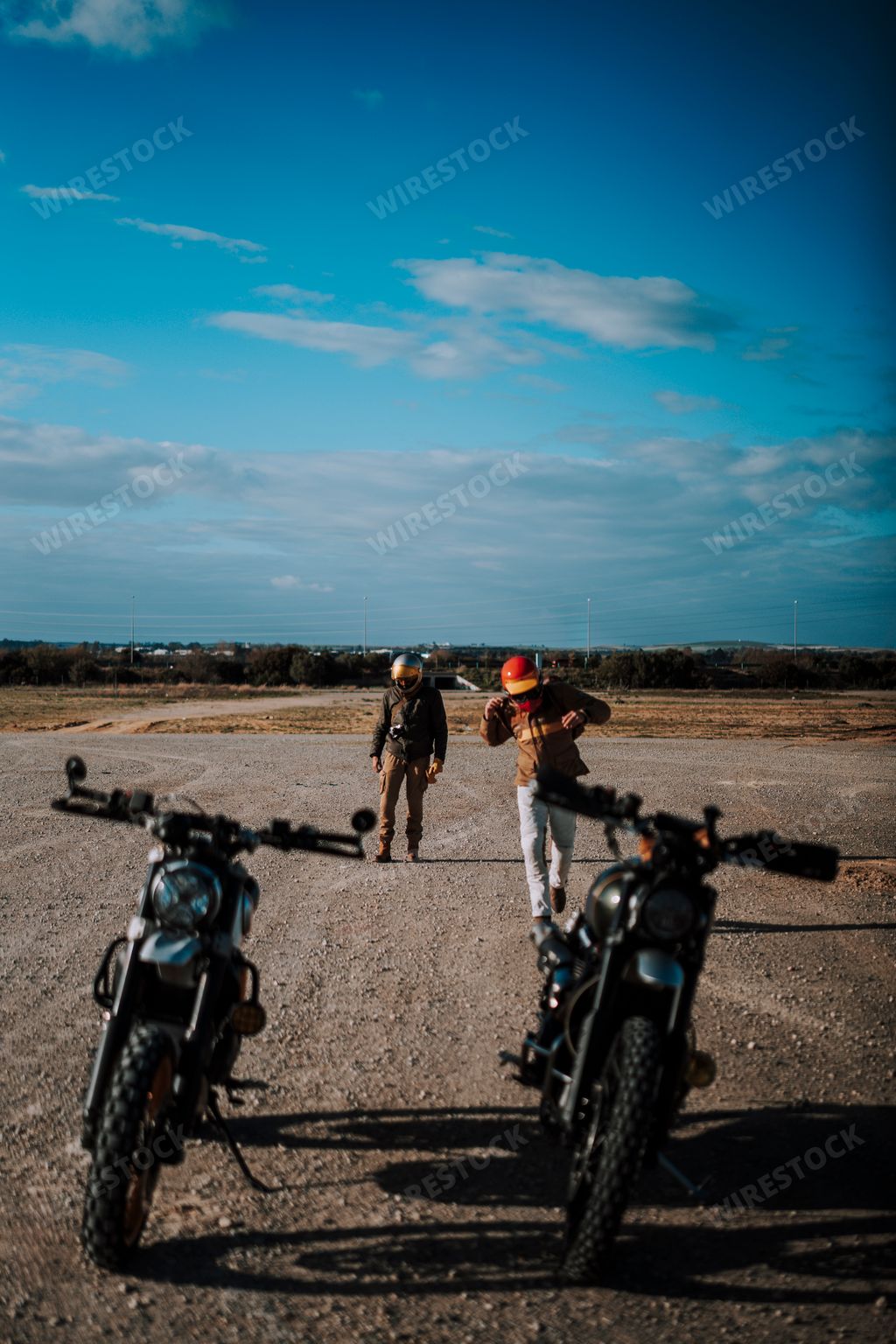 Two individuals stroll down a desert road, with motorcycles parked on either side
