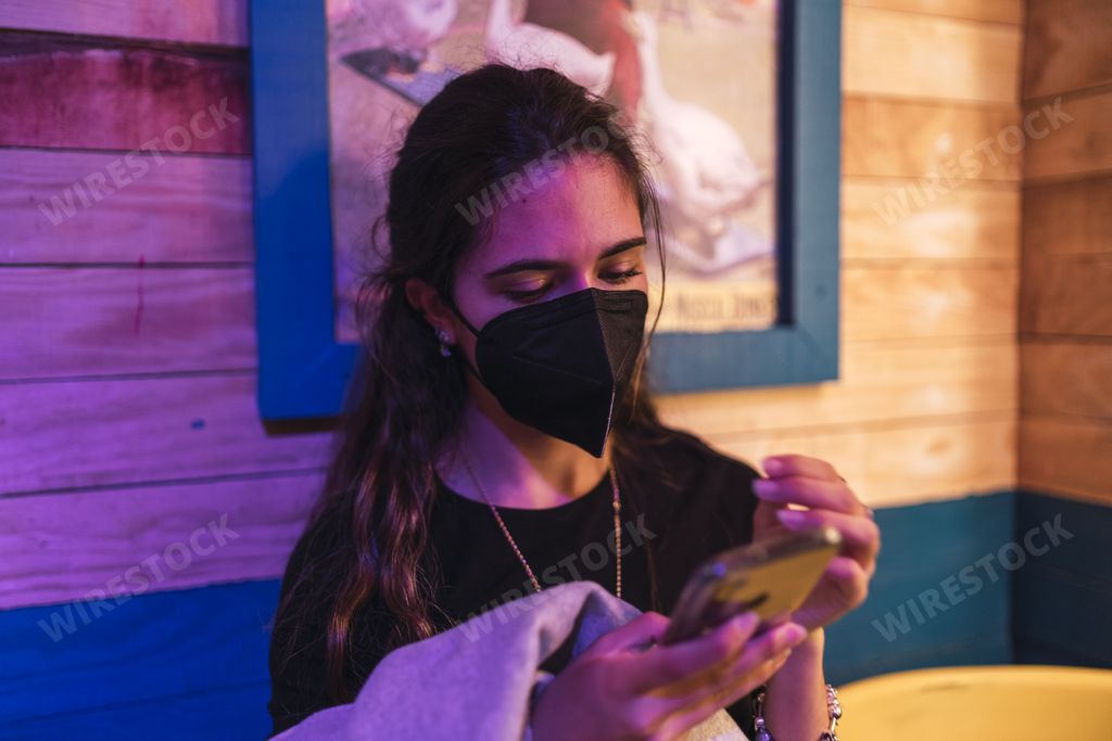 A young Spanish girl wearing a black face mask is scrolling on the smartphone while resting in a cafe