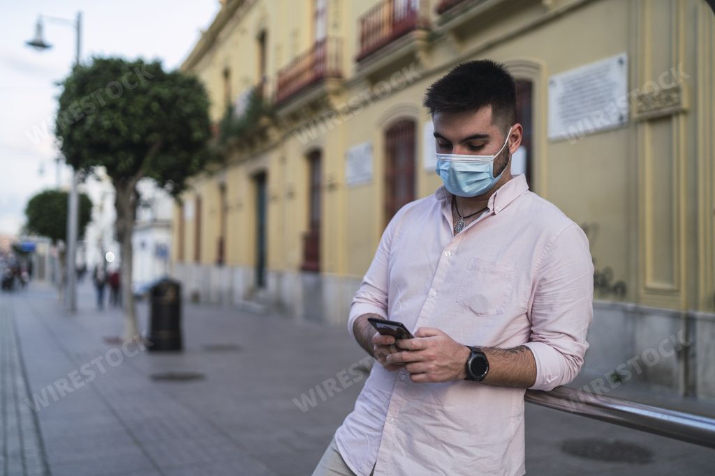 A handsome Spanish man wearing a mask and typing on his phone - new normal concept