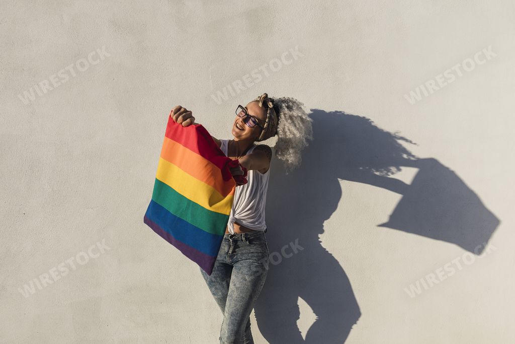 A curly-haired Spanish cute woman with a pride flag tote bag and tattoos on a white wall background