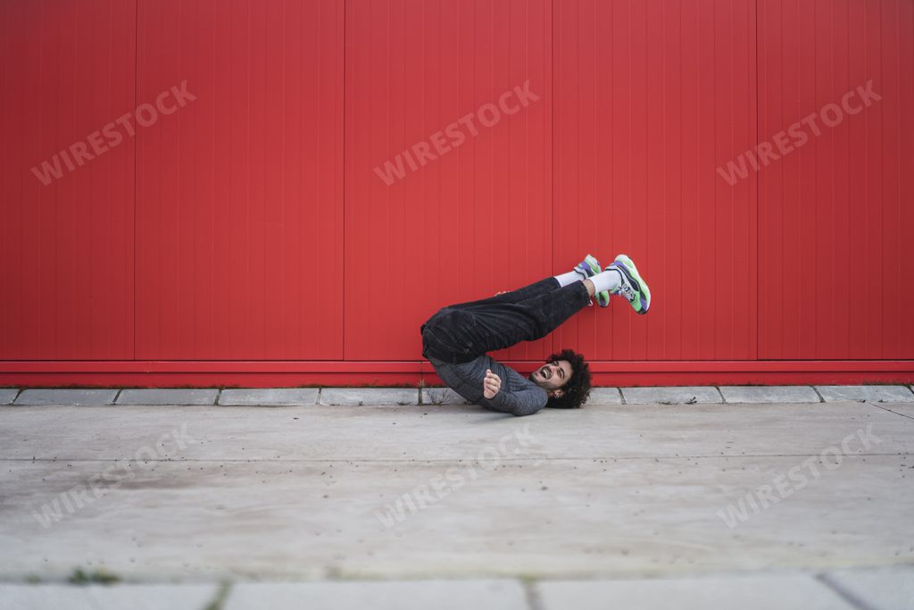 A laughing Spanish guy with curly hair laying on the asphalt and doing a reverse crunch in the red wall background