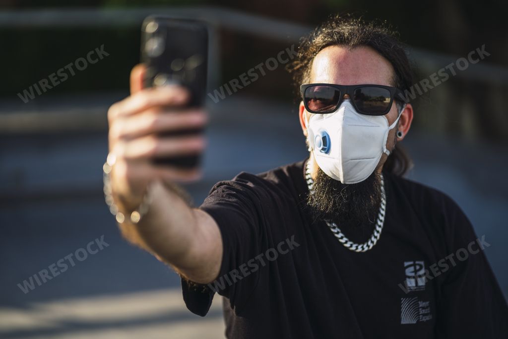 A selective focus shot of a bearded male in a black shirt wearing a medical face mask while taking a selfie from his phone- Covid-19 concept