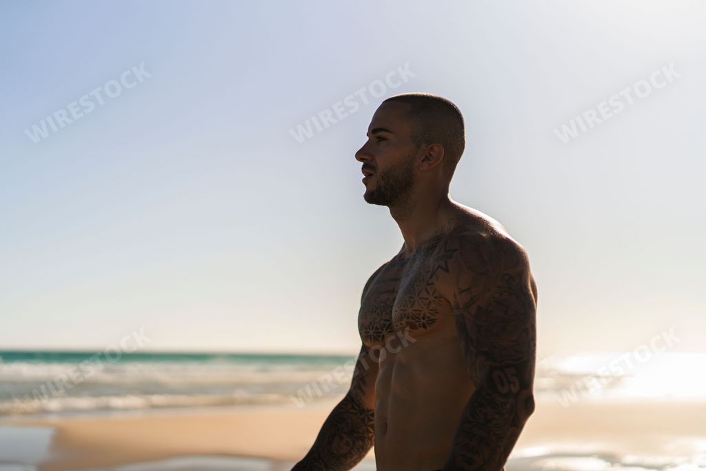 A closeup shot of an attractive topless man with tattoos standing  on a beach next to a sea