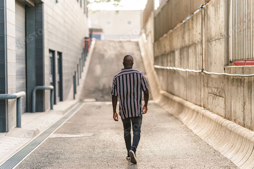 A back view of a fit black male with a striped shirt and jeans walking in an alley